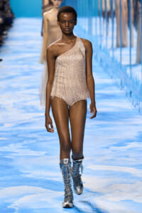 Model walking a runway in a one-shoulder, champagne-colored sequined bodysuit with metallic lace-up boots on a blue-toned catwalk