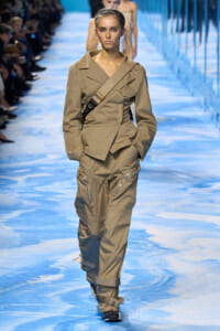 Model walks the runway in a tan utility suit with a crossbody strap and zip-pocket pants.