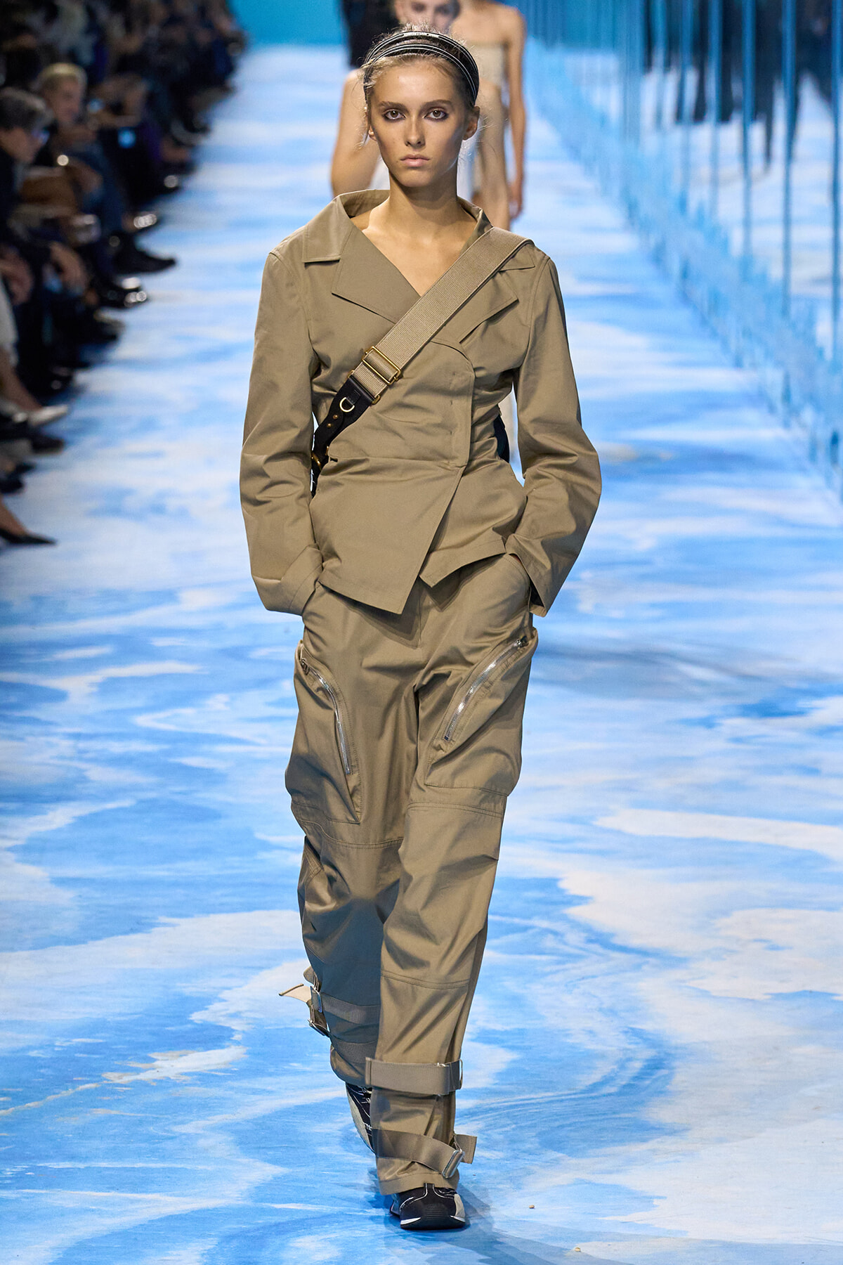 Model walks the runway in a tan utility suit with a crossbody strap and zip-pocket pants.