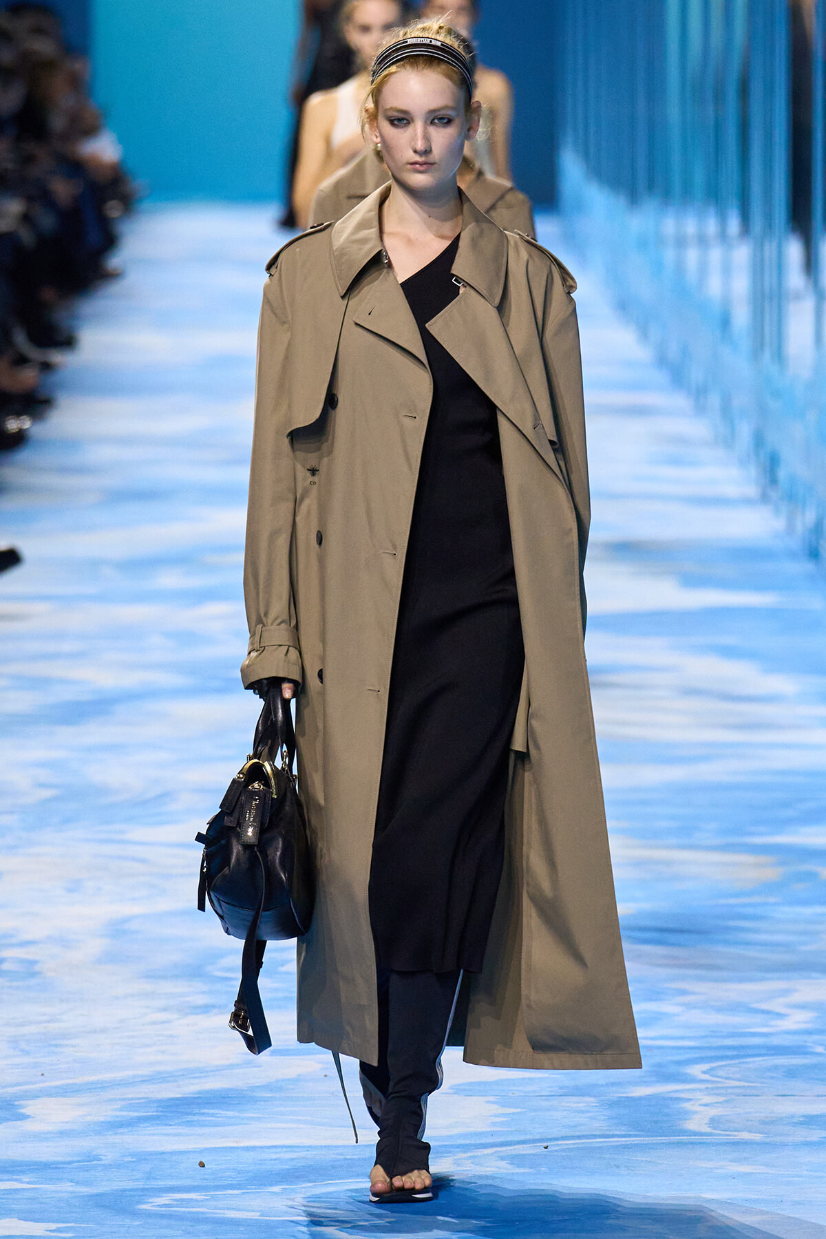Model walks a fashion runway wearing a tan trench coat over a black dress, carrying a dark handbag.