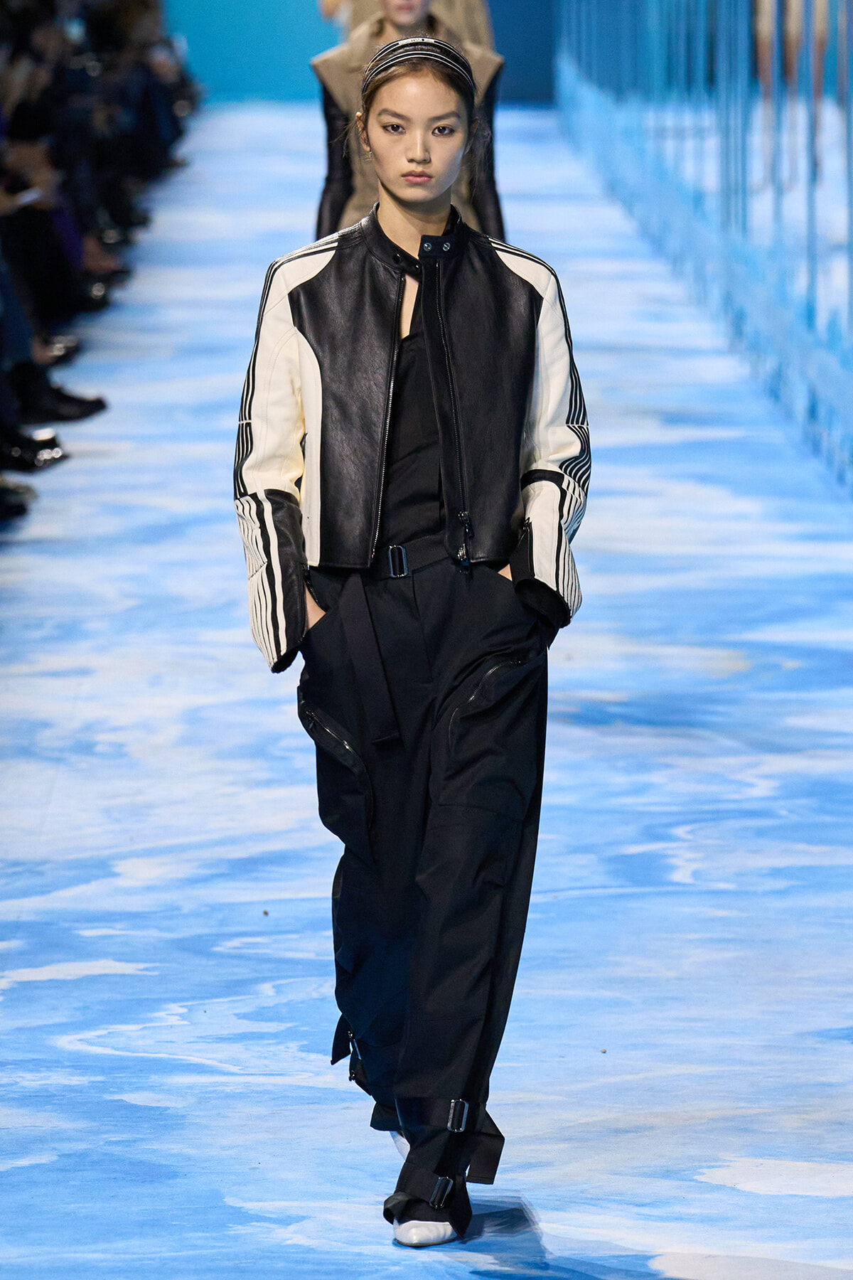Female runway model wears a black-and-white leather jacket and black cargo pants on a blue runway.