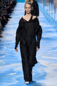Model walking a blue runway in a black oversized jumpsuit with exposed shoulders and zippers.