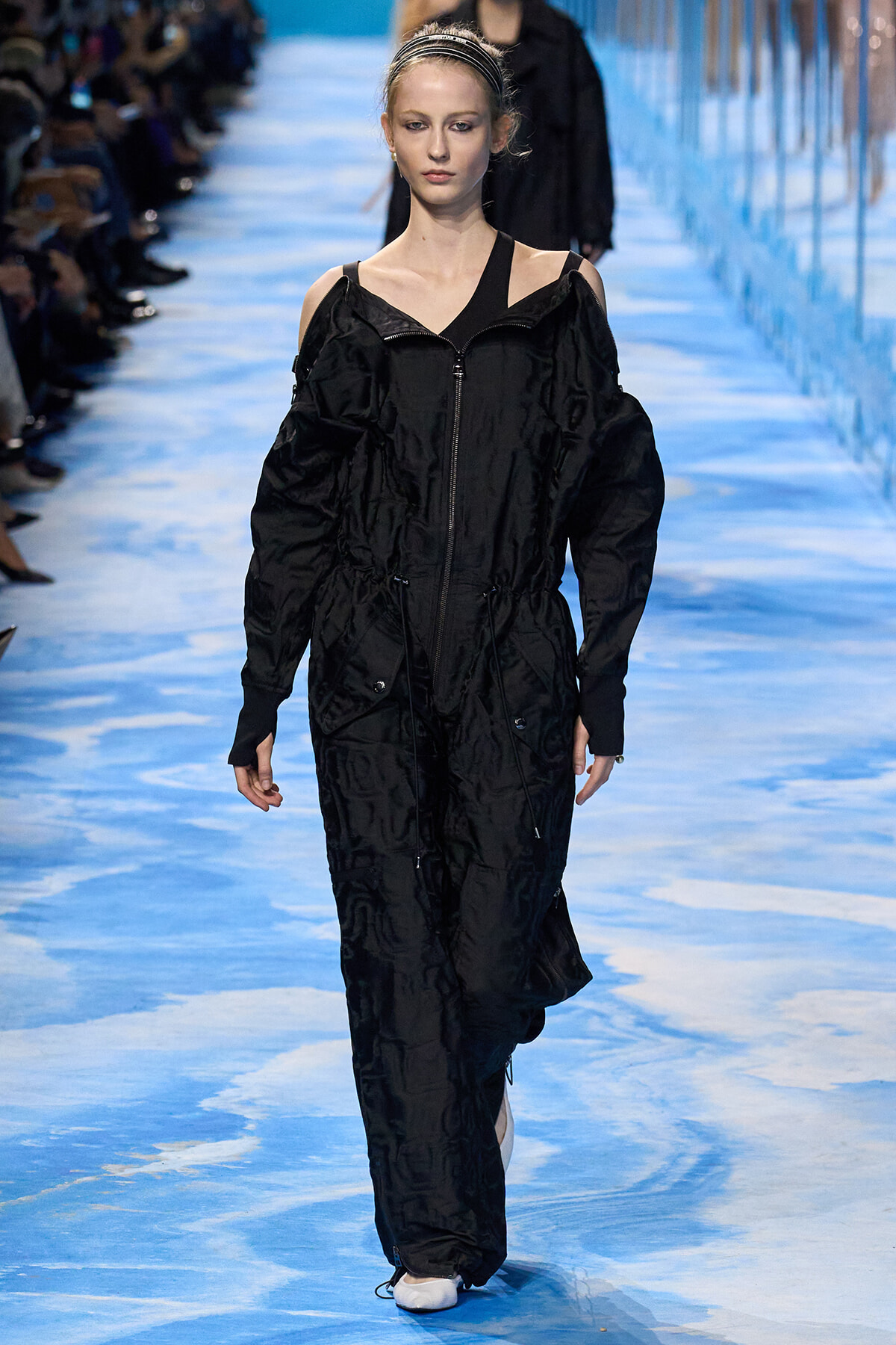 Model walking a blue runway in a black oversized jumpsuit with exposed shoulders and zippers.