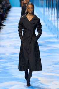 Model walks the runway in a long black trench coat with ruched sleeves and black gloves on a blue-toned catwalk.
