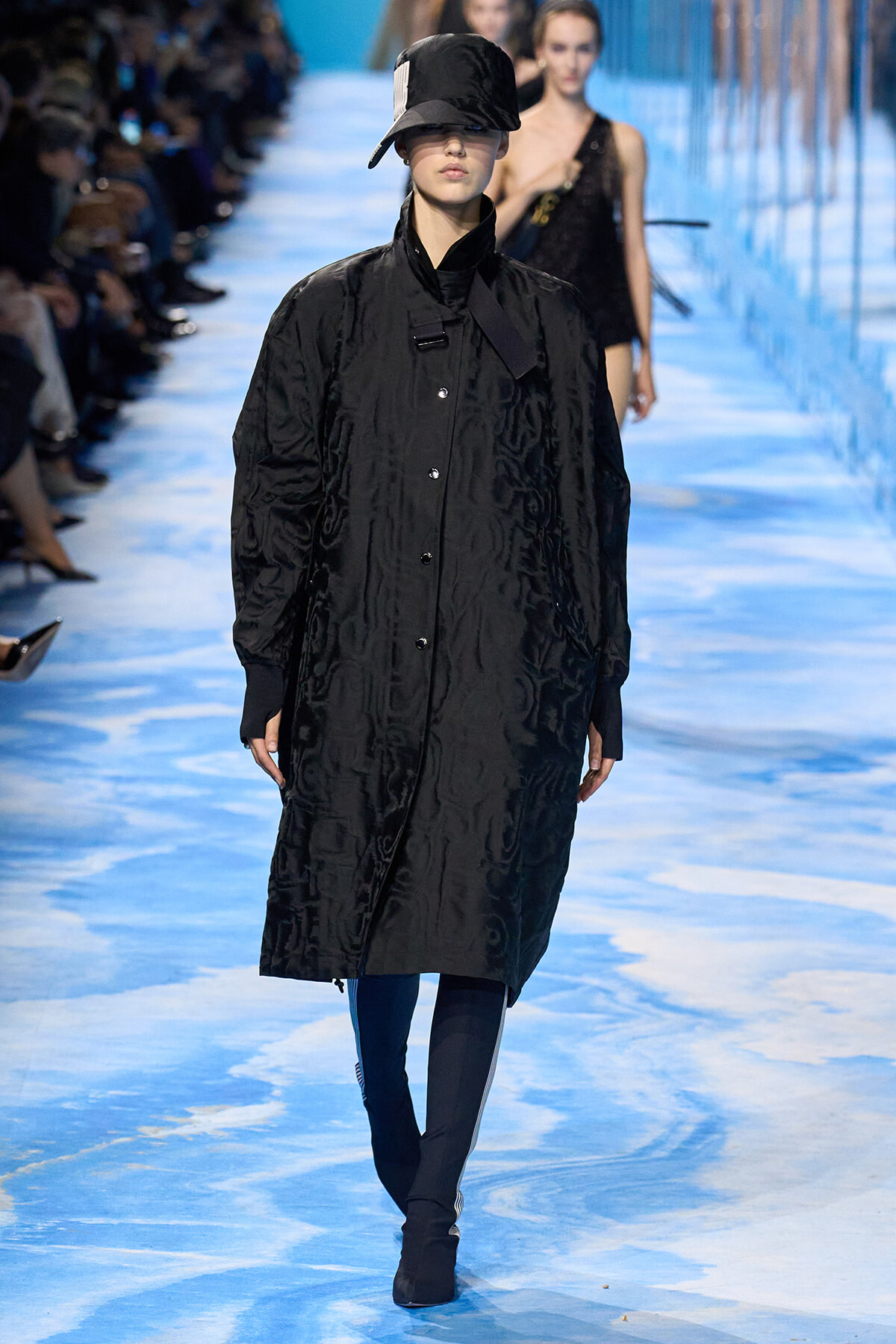 Model on a blue runway wearing a long black coat with a tilted cap and blue tights.