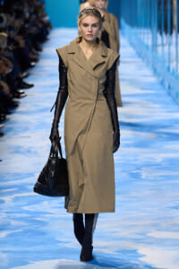 Model walks the runway in a tan sleeveless trench dress with black long gloves, headband, and a black quilted handbag.