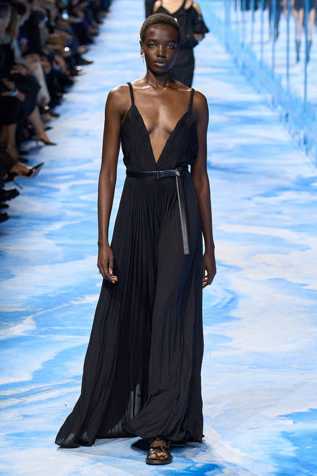 Model walks the runway in a black sleeveless pleated gown with a deep V-neck and a thin belt.