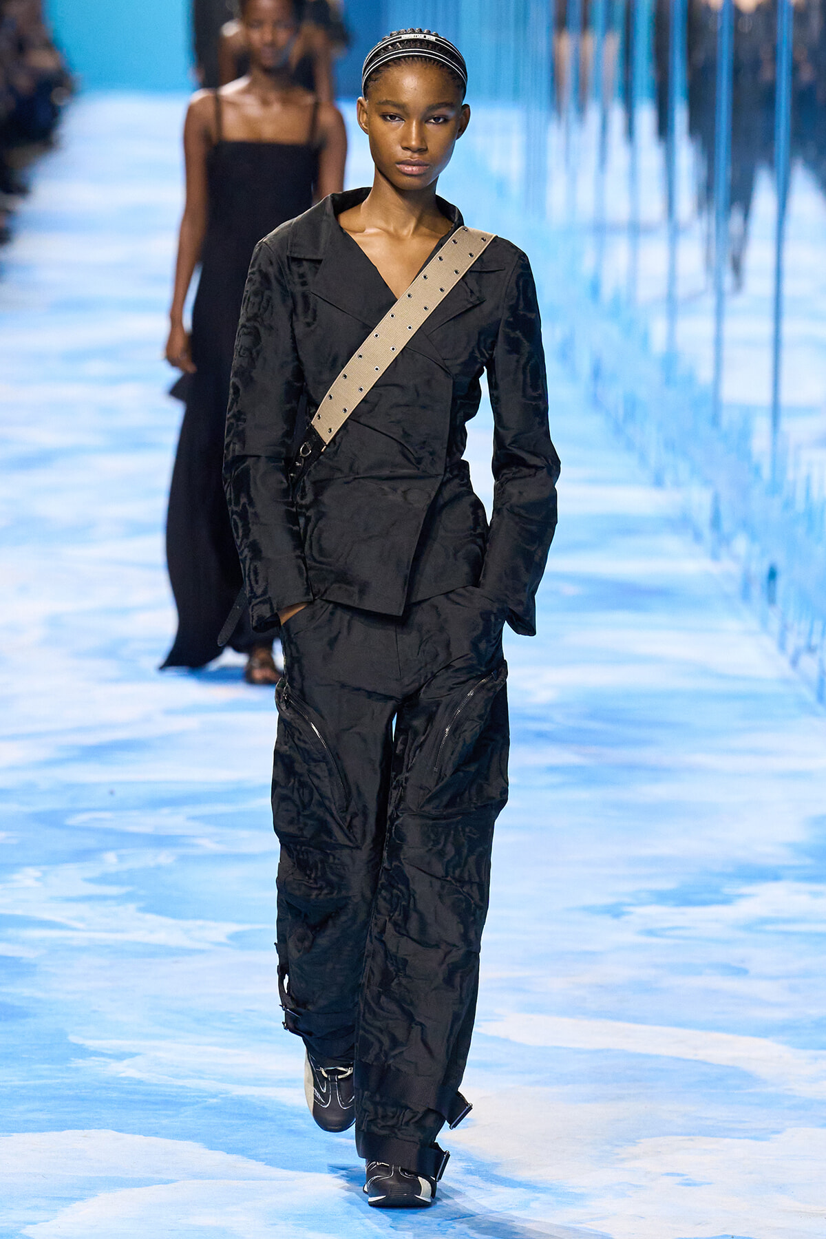 Model walks the runway in a black utilitarian jumpsuit with a beige diagonal crossbody strap on a blue-lit catwalk