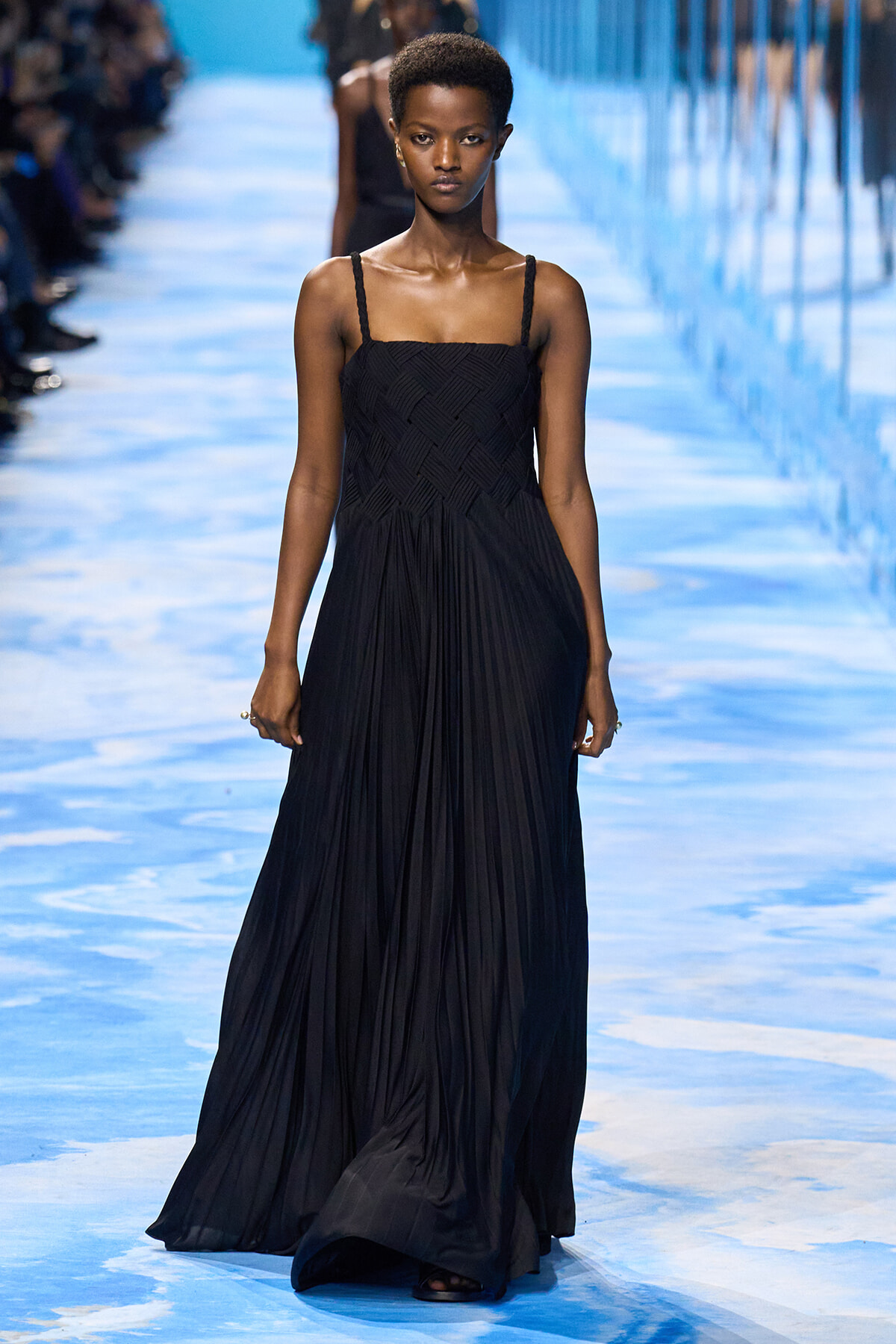 African model walks down a blue-lit runway wearing a long black pleated gown with thin straps and a fitted bodice on a fashion show catwalk.