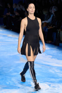 Runway model wearing a black sleeveless top and a high-waisted skirt with slit panels, paired with black knee-high boots with white stripes.