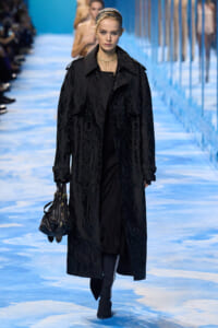 Female model walking a fashion show runway in a long black patterned trench, layered pearls, and a black handbag.