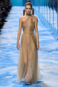 Model walks the runway in a beige, sheer, halter-neck gown with a wrapped bodice and flowing overlay on a blue-patterned runway.