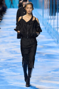 Female model walking a blue-lit runway in a black off-shoulder windbreaker with zipped front and matching pants, hands in pockets.