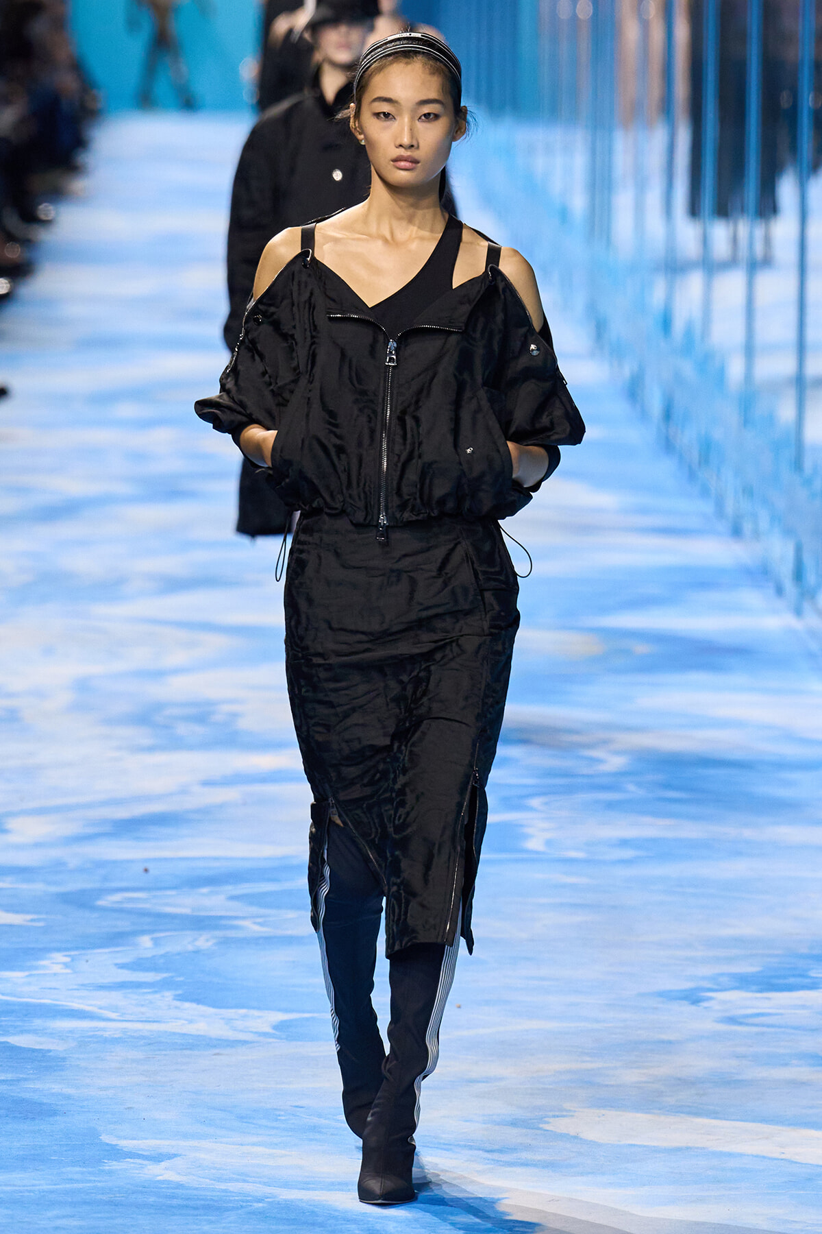 Female model walking a blue-lit runway in a black off-shoulder windbreaker with zipped front and matching pants, hands in pockets.
