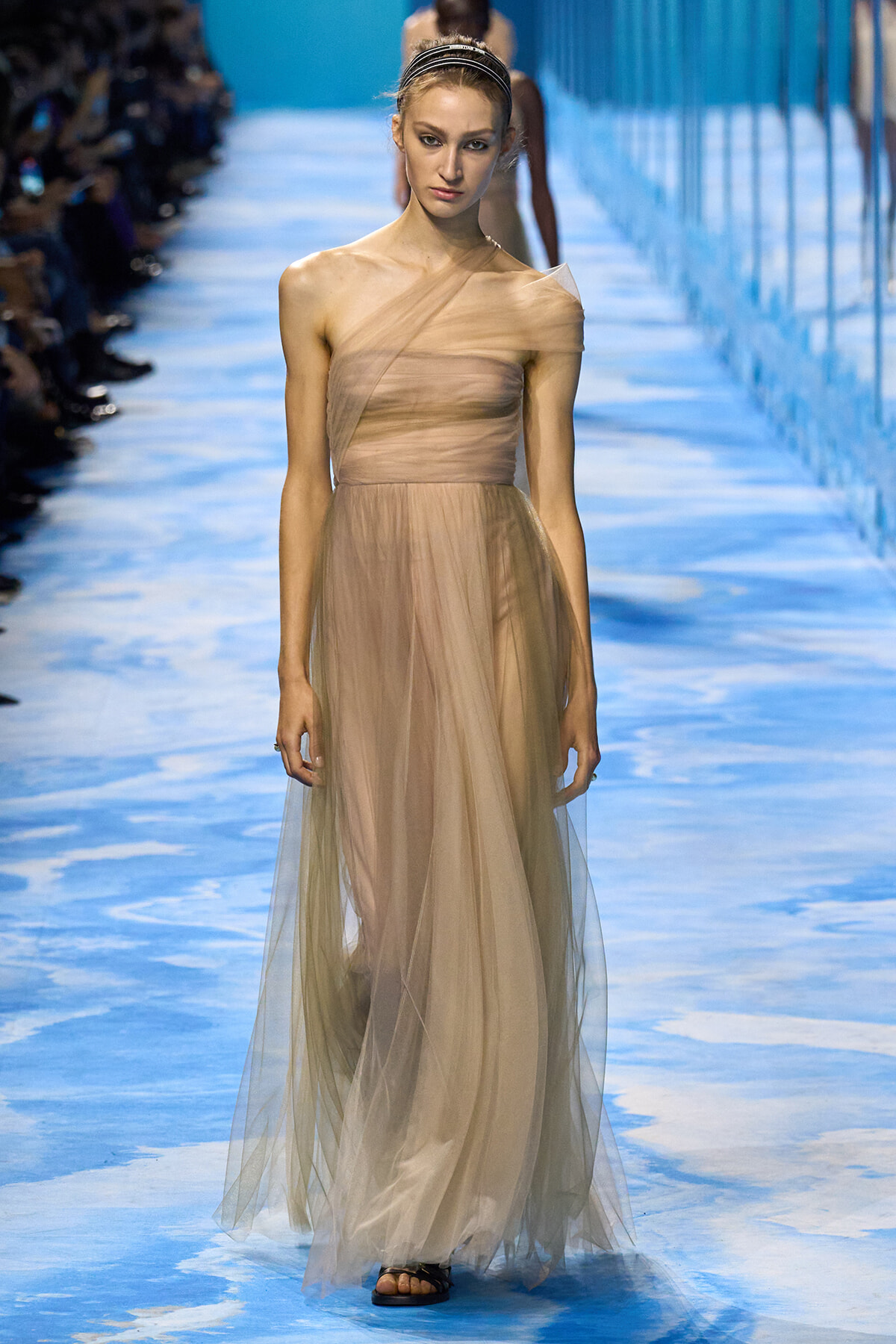 Model walking a runway in a nude-toned, one-shoulder tulle gown with a sheer overlay and headband, on a blue patterned catwalk