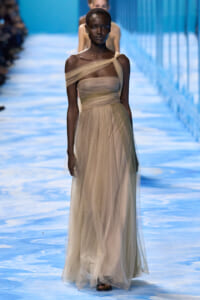 Model walks a blue-tinted runway wearing a beige one-shoulder tulle gown with a draped bodice and sheer skirt.