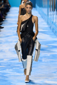 Model walks the runway in a black one-shoulder top, black leather trousers, and a white‑black geometric coat draped around her hips on a blue runway backdrop.