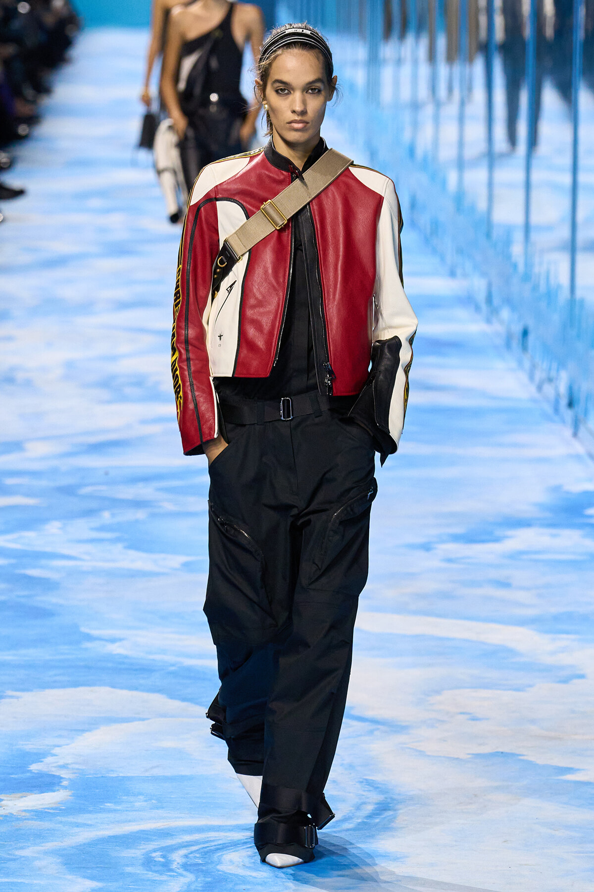 Model walks the runway in a color-block red, white, and black leather jacket with a beige crossbody strap over black cargo pants on a blue-toned catwalk