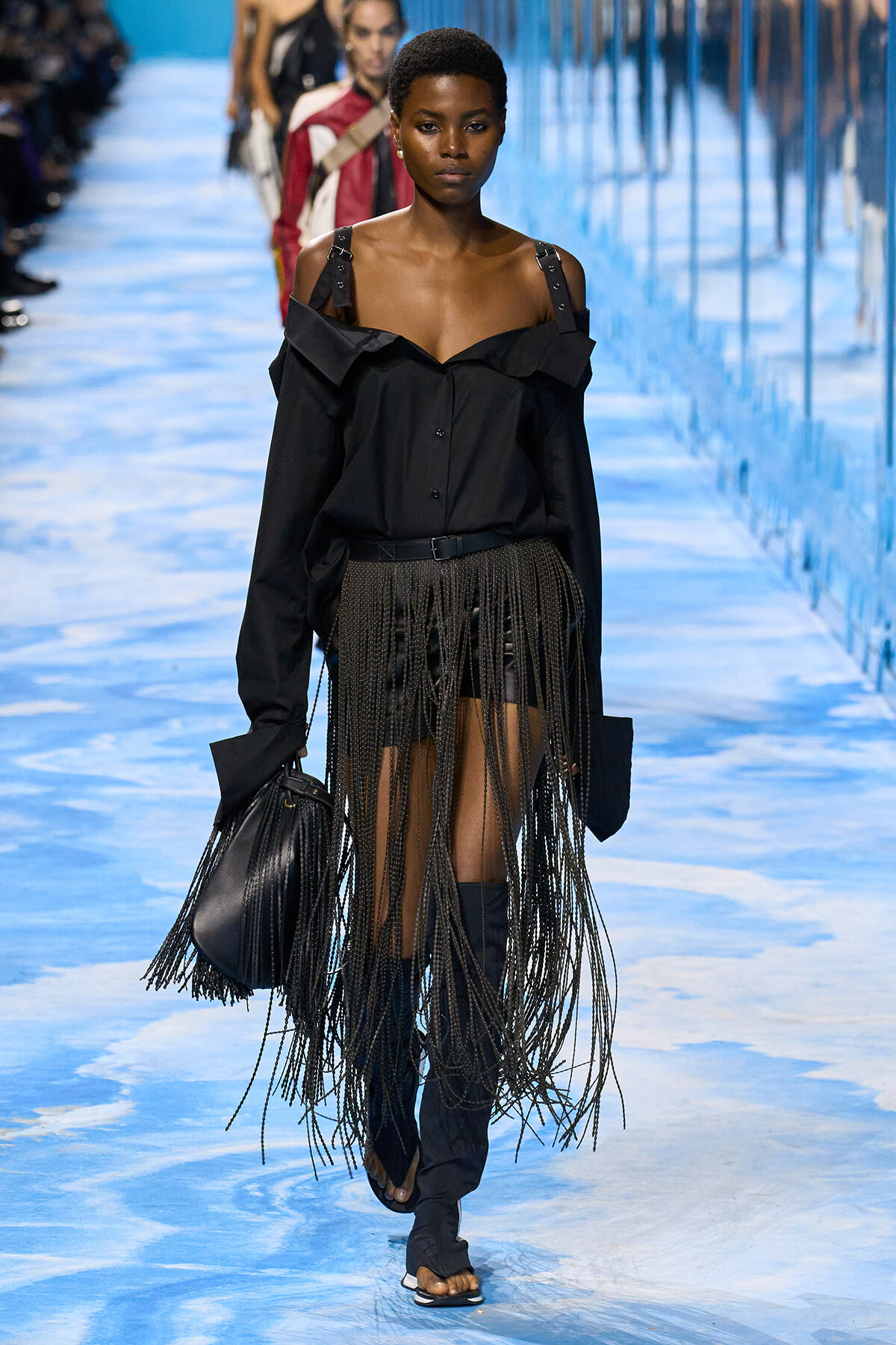 Model walking a fashion show wearing an off-shoulder black blouse with a fringe-detail skirt and knee-high boots, carrying a black bag.