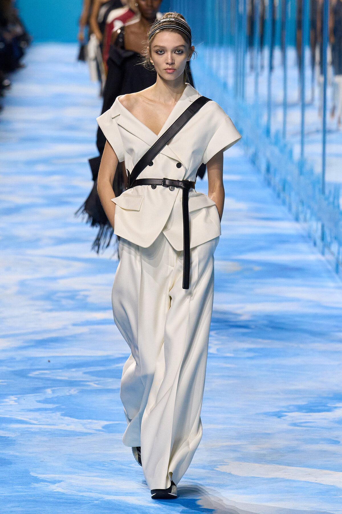 Model walks the runway in a white oversized suit with a black belt and crossbody strap, hands in pockets.