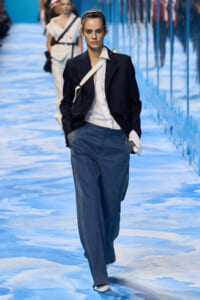 Model walks the runway in a navy blazer, white shirt, and loose blue trousers with a white crossbody bag and headband on a blue reflective catwalk.