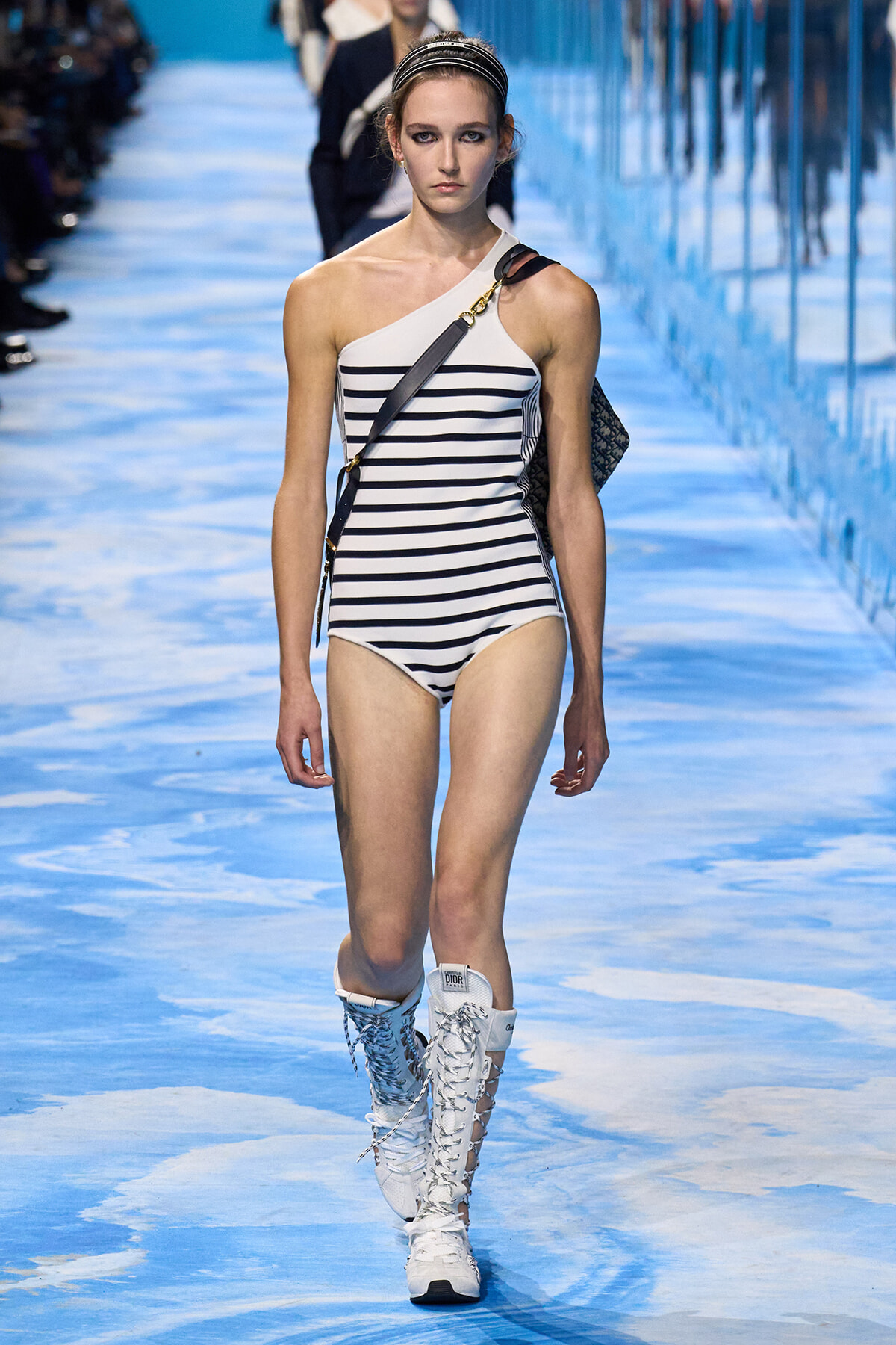 Model walks the runway in a white and navy striped one‑shoulder swimsuit with a crossbody bag and white lace‑up boots.