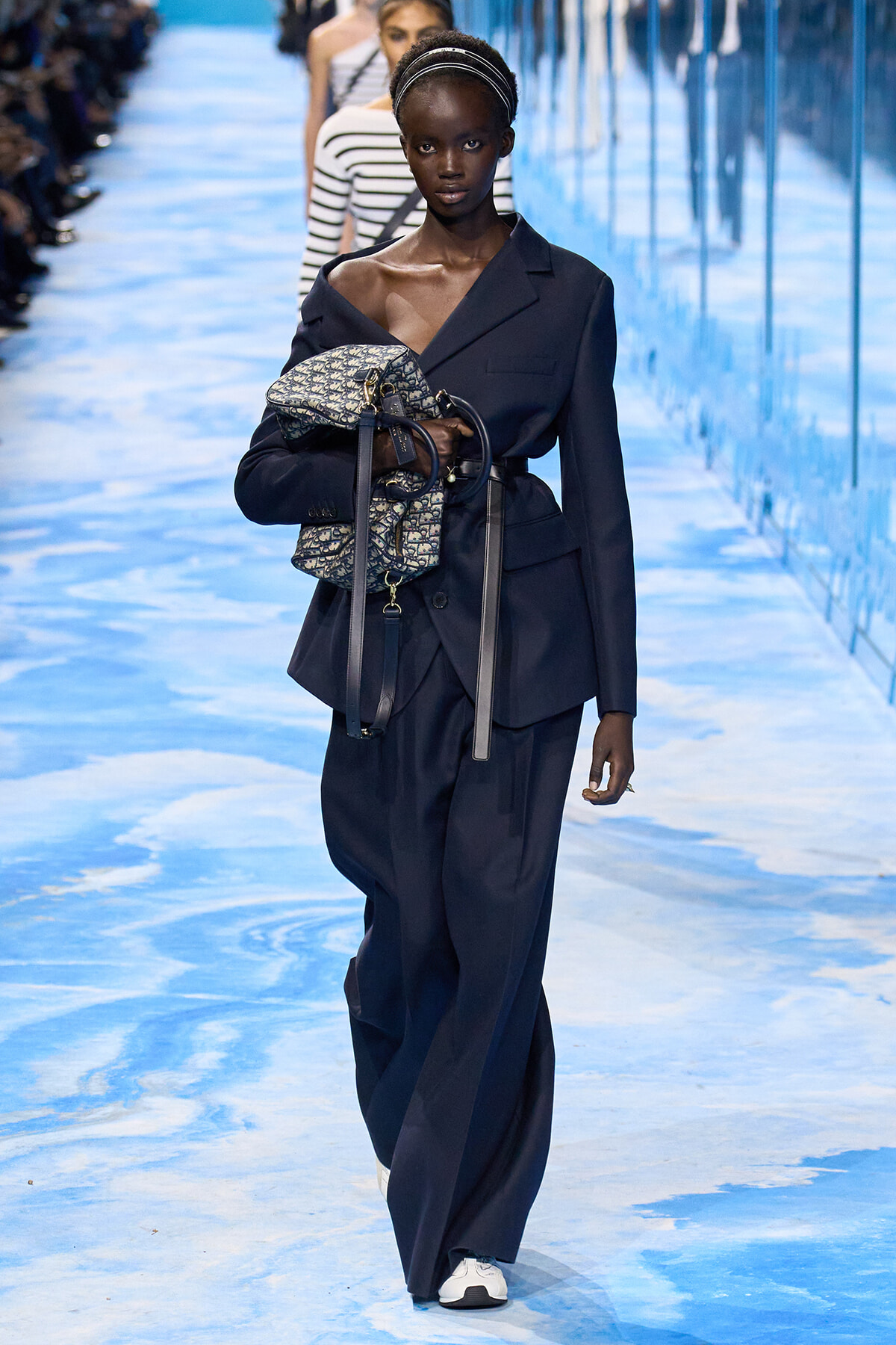 Model walks a blue-toned runway in a navy oversized blazer and wide-leg pants, clutching a patterned designer backpack with dark straps.