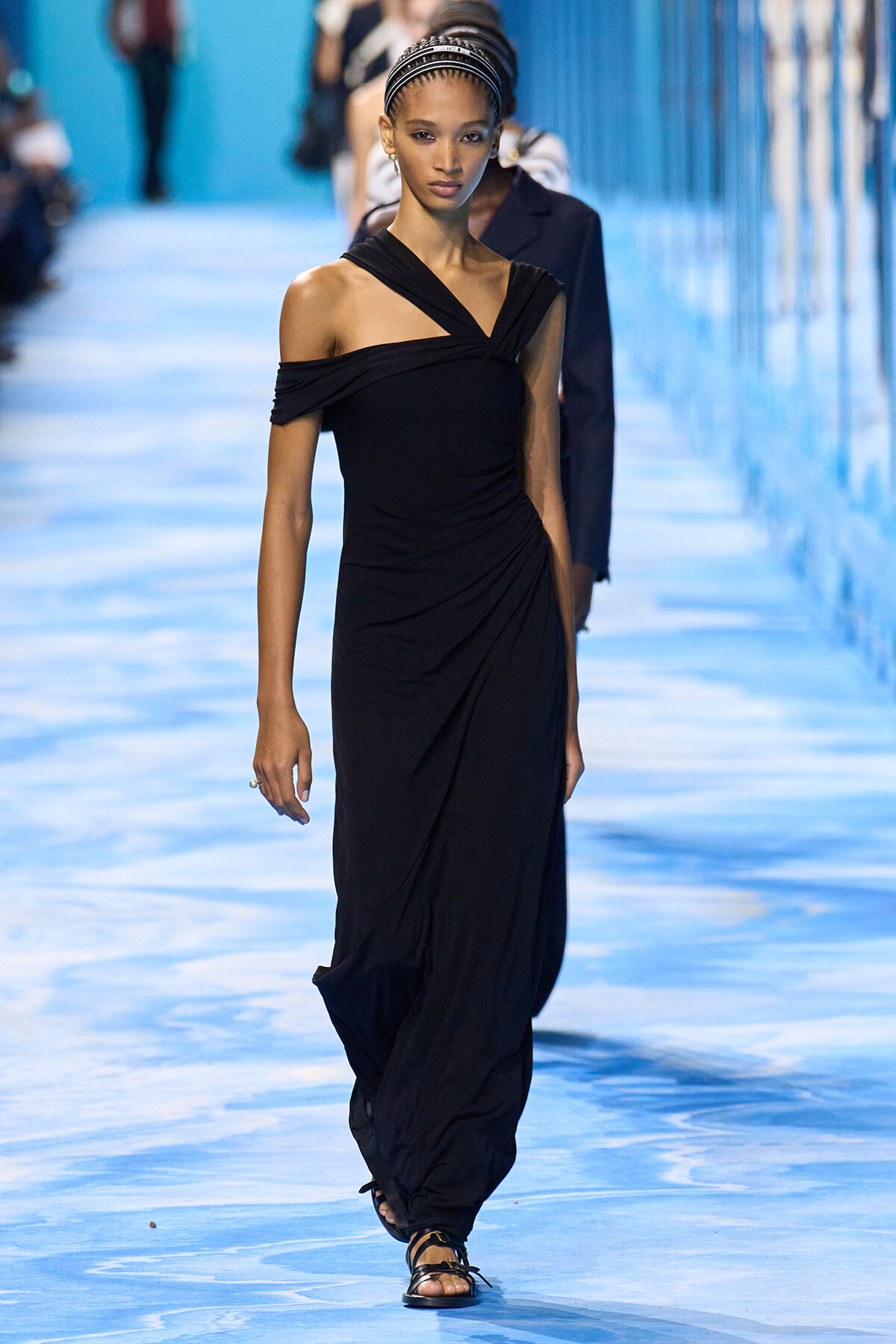 Model walking a blue-toned runway in a black off-shoulder, twisted-strap gown with draped waist.
