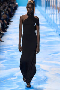 Model walks a blue-patterned runway in a long black one-shoulder gown with a crisscross strap across the chest, wearing sandals.