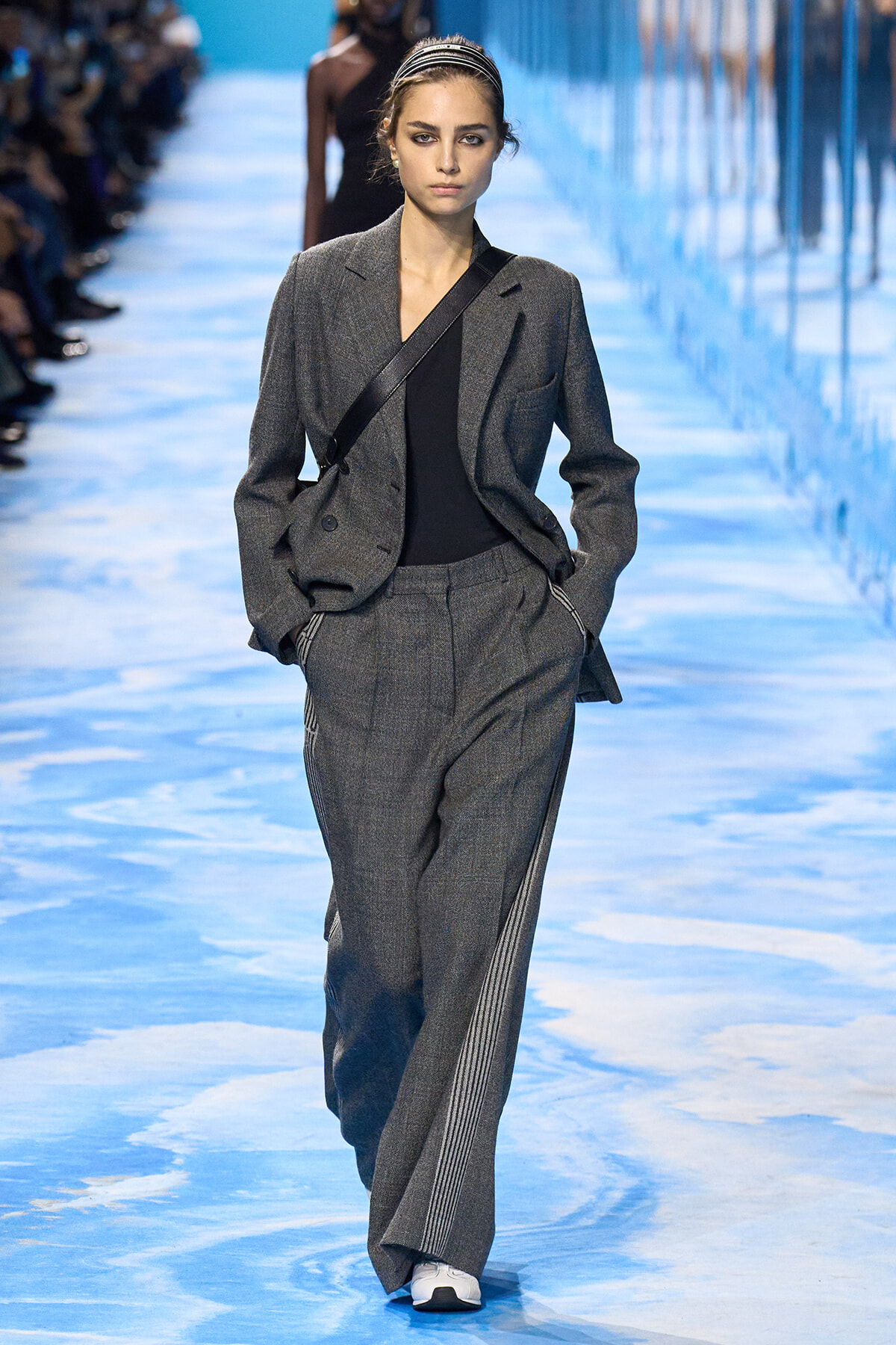 Model walking a fashion runway in a gray suit with a black top, crossbody bag, and white sneakers against a blue-lit backdrop