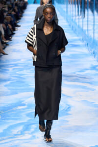 Model walking the runway in a black double-breasted jacket with a striped white sleeve detail, paired with a long black skirt and sandals.