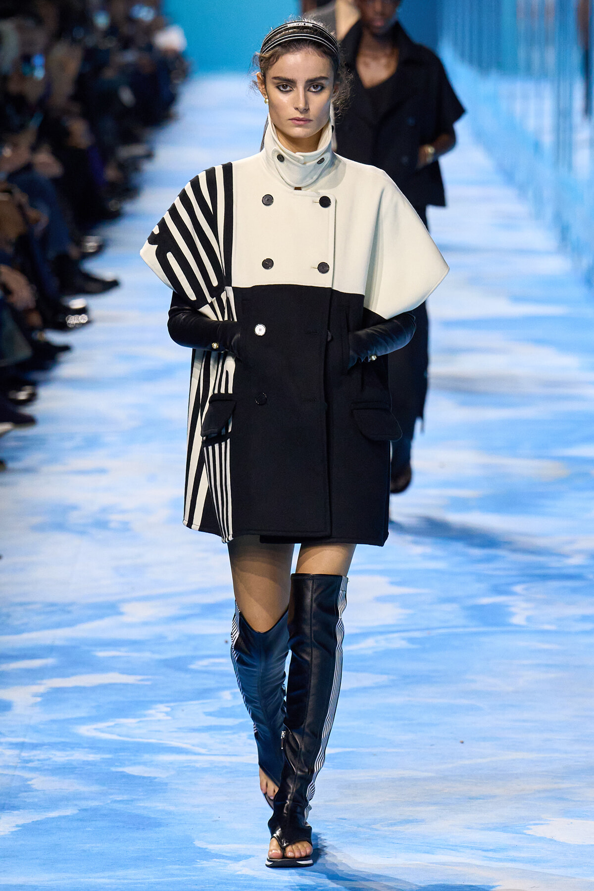 Model walks the runway in a black-and-white color-block coat with bold striped cape detail and knee-high black boots on a blue-tinted catwalk.