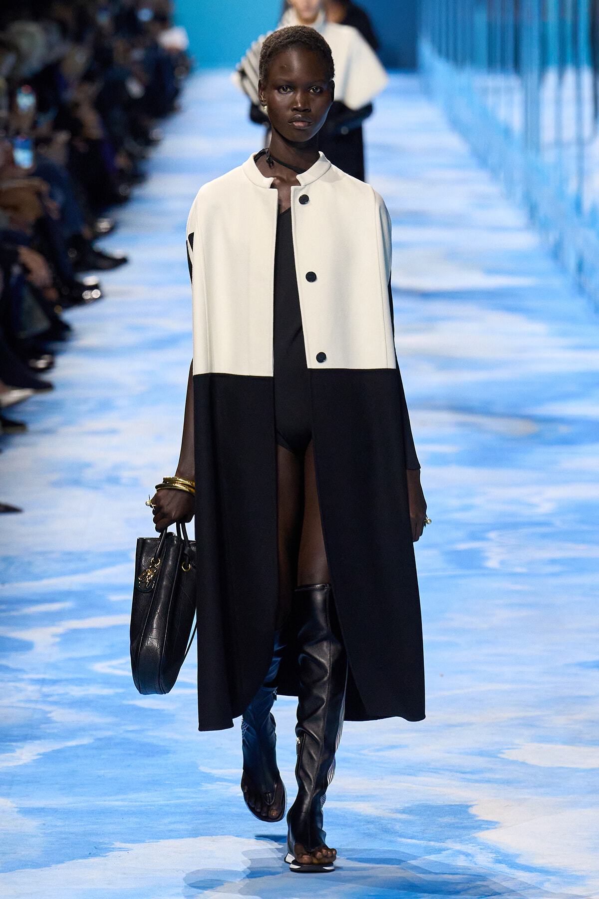 Model walks a blue runway wearing a two-tone white-and-black midlength coat over a black outfit, carrying a black handbag and wearing knee-high boots.