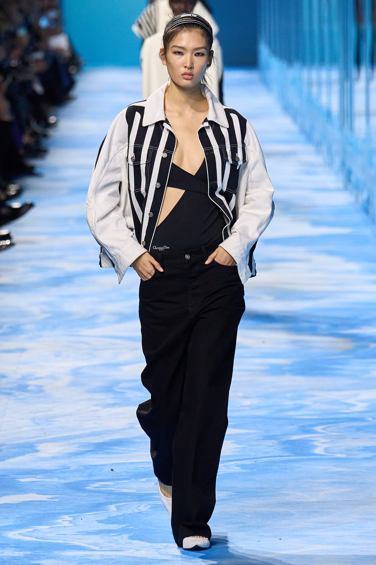 Model walks the runway in a white and black striped jacket over a black cutout top and wide-leg black pants on a blue catwalk.