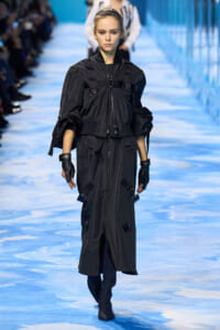 Model walking a runway in a black oversized utility coat with straps and pockets, gloves, and knee-high boots.