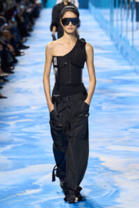 Female model walks the runway in a black one-shoulder top with harness details and baggy cargo pants, wearing oversized sunglasses.