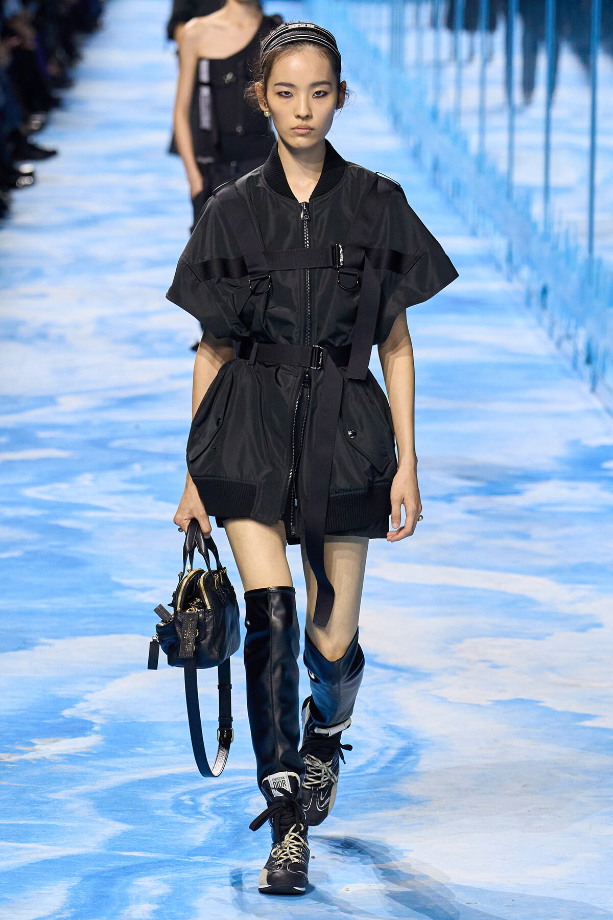 Model walks the runway in a black utilitarian jacket with belt, matching shorts, and tall black boots, carrying a small handbag.