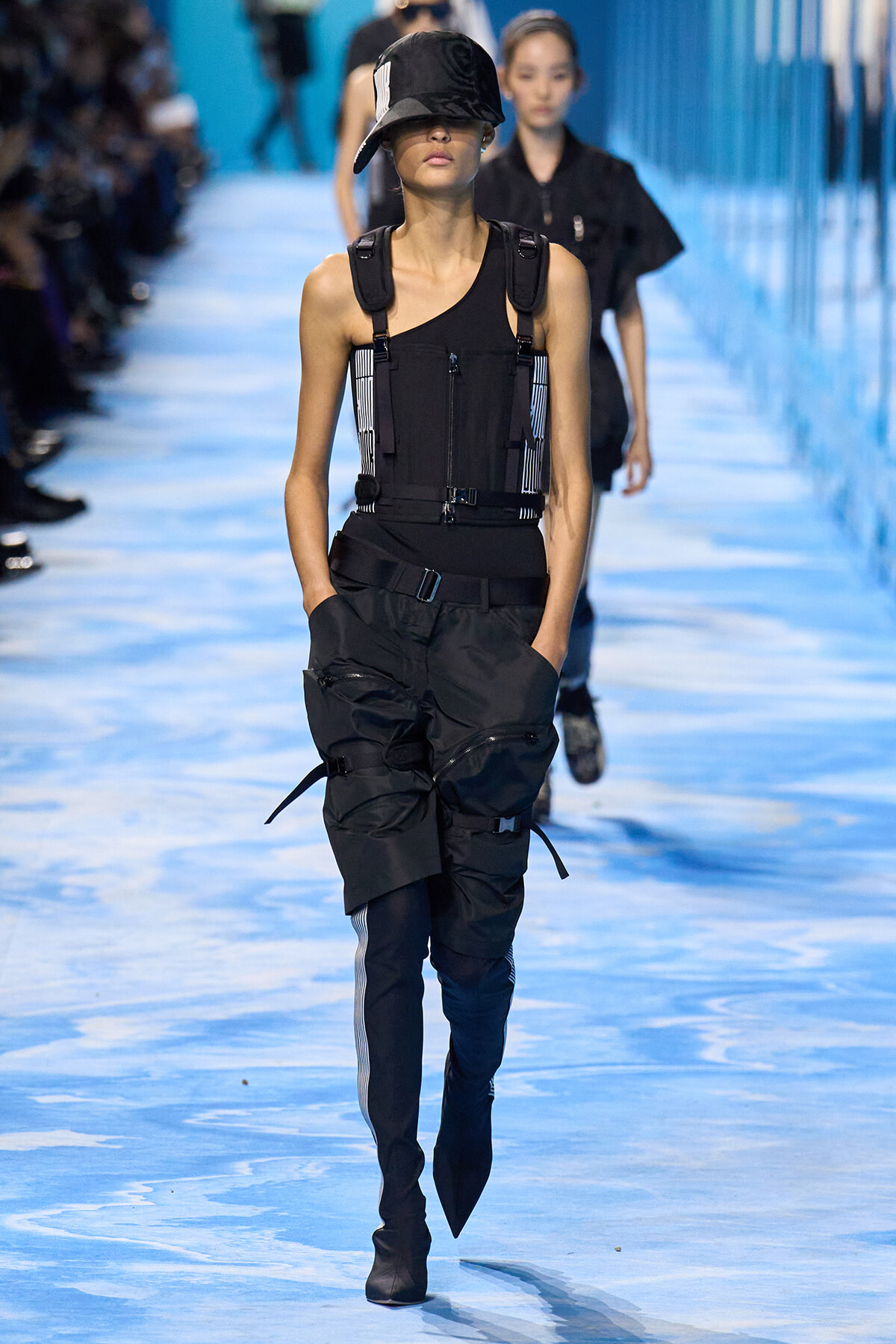Model on a blue runway wearing a black utilitarian outfit with harness straps and oversized cargo pants, hands in pockets.