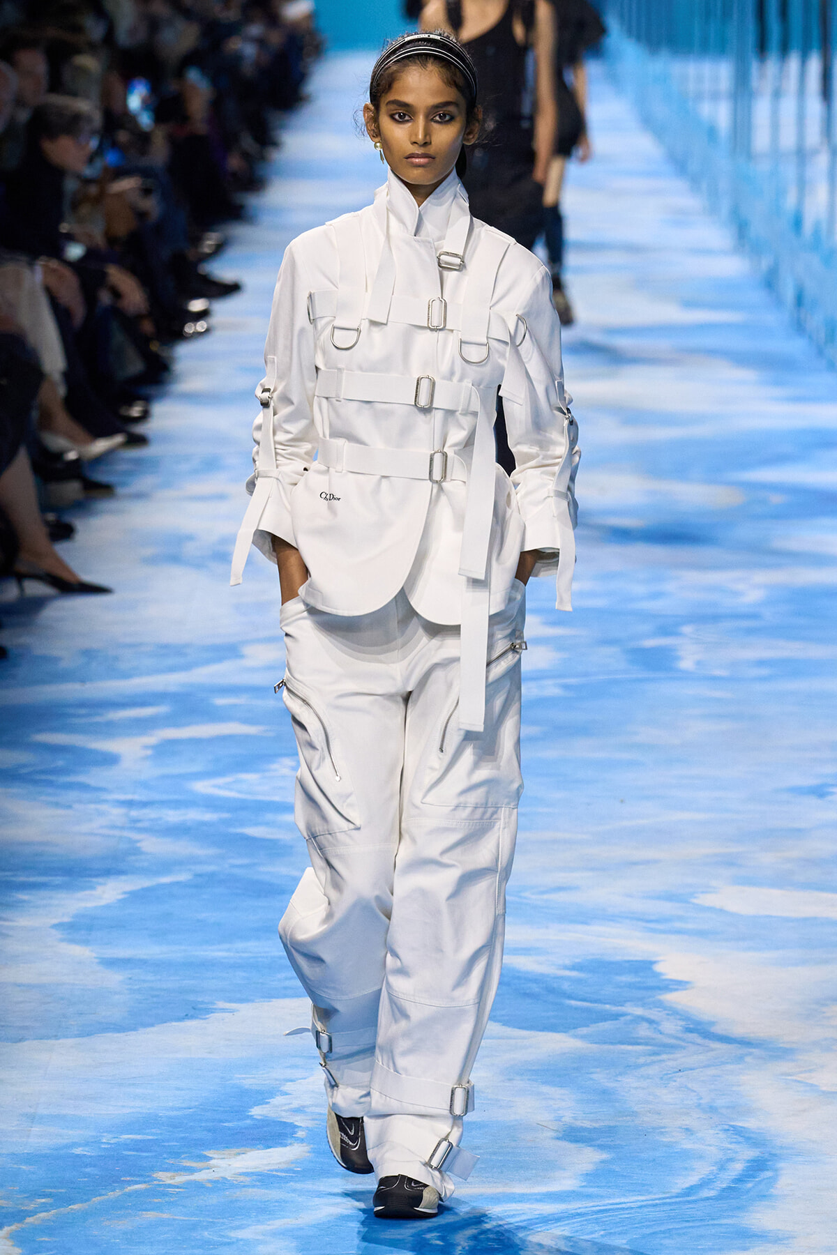 Runway model in a white harness-style coat with multiple buckles and white cargo pants on a blue-toned catwalk.