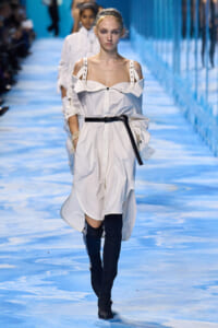 Model walks a fashion runway in a white off-shoulder shirt dress with a black belt and black thigh-high boots on a blue-toned catwalk.