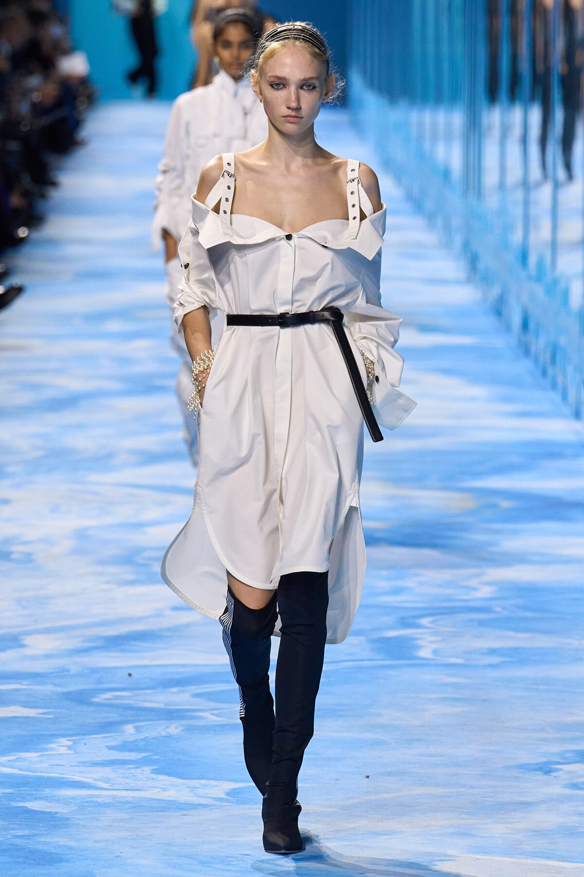 Model walks a fashion runway in a white off-shoulder shirt dress with a black belt and black thigh-high boots on a blue-toned catwalk.