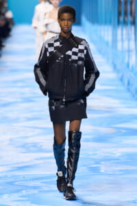 Model on a blue-lit runway wearing a black jacket with white checkered panels, crossbody strap, and a black mini skirt with tall blue and black boots.