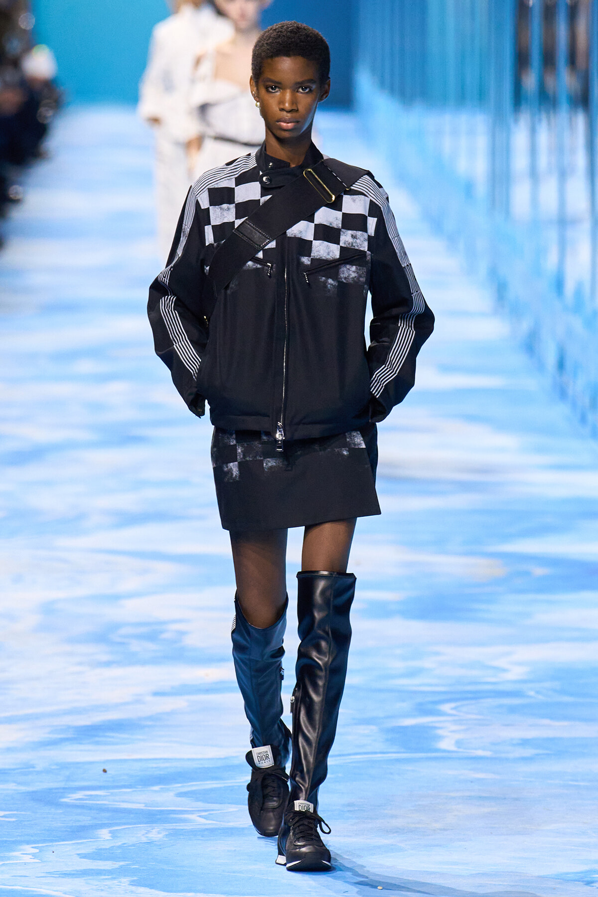 Model on a blue-lit runway wearing a black jacket with white checkered panels, crossbody strap, and a black mini skirt with tall blue and black boots.