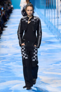 Model walking the runway in a black and white checker jacket and pants on a blue-lit catwalk.
