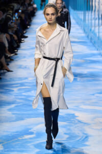 Model walking a fashion runway in a white shirt dress with a black belt and knee-high black boots along a blue-toned catwalk.