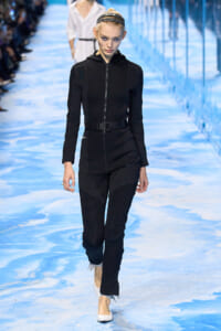 Model walking a blue runway in a black zip-front jumpsuit with a belt and white flats, hair tied back with a headband.