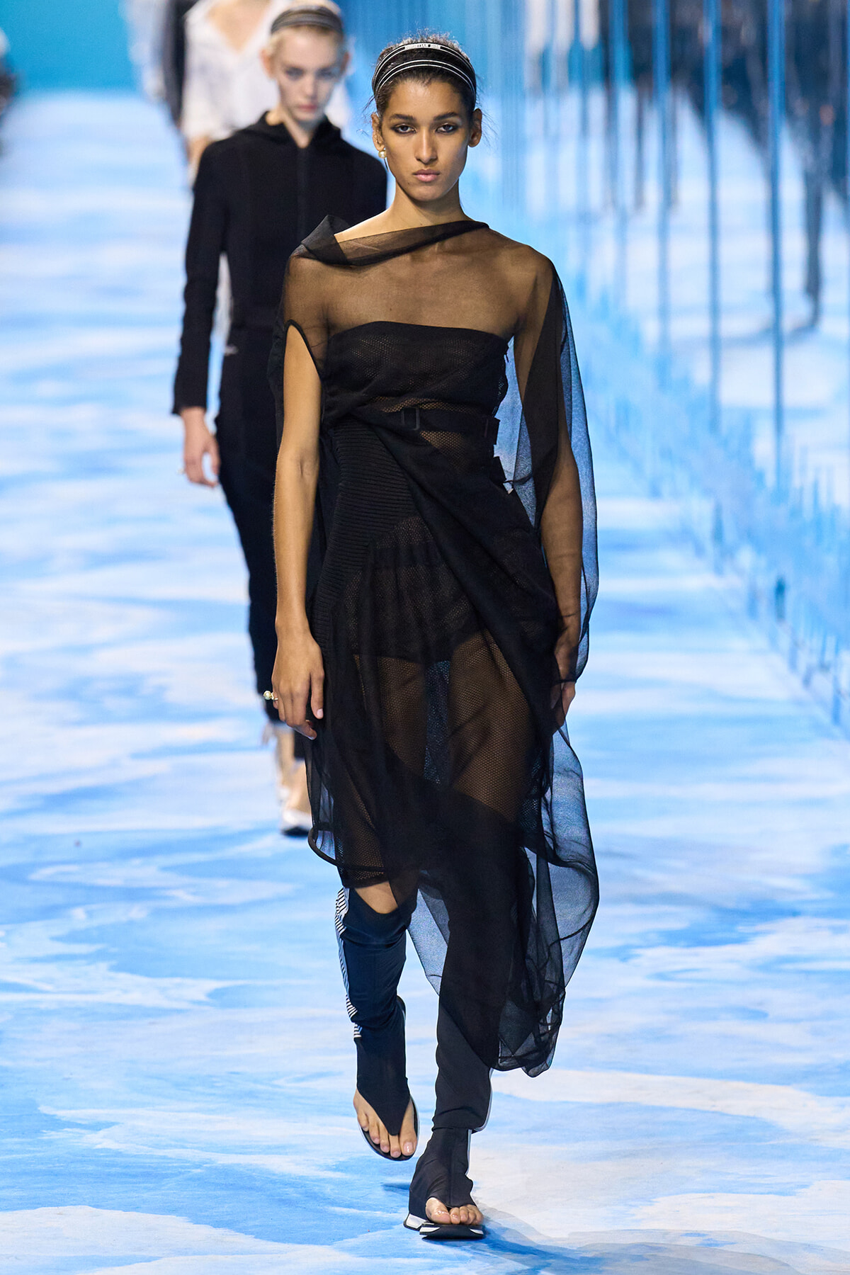 Model on a runway wearing a sheer black draped dress with a belt, headband, and wrap-style sleeves against a blue-toned background.