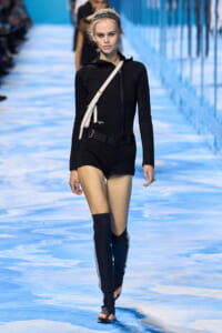 Model walks the runway in a black zip-front bodysuit, black belt, and knee-high black boots with a white crossbody bag over one shoulder on a blue-patterned runway.