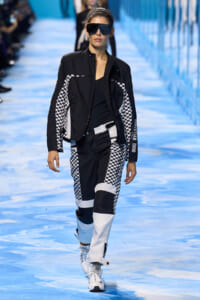 Model walks a blue runway in a black and white checkerboard racing-inspired outfit, oversized black jacket, and white sneakers plus large sunglasses.