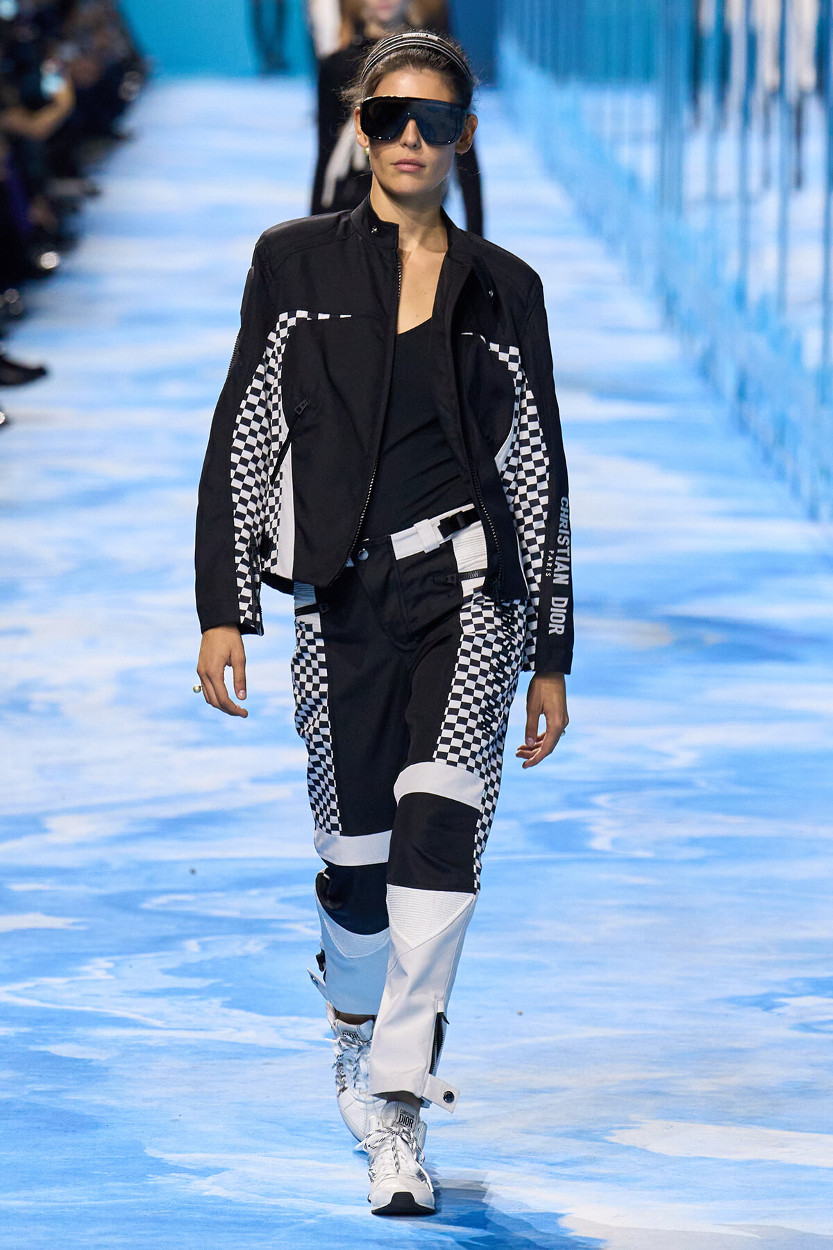 Model walks a blue runway in a black and white checkerboard racing-inspired outfit, oversized black jacket, and white sneakers plus large sunglasses.